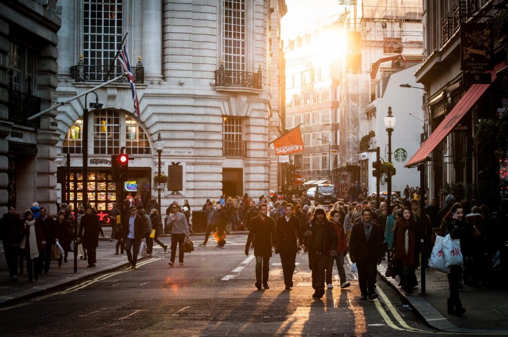 photo of potential scene of a pedestrian accident featured by pedestrian accident lawyer david buckley, features pedestrians walking in a busy street, a sun setting between two buildings in the right third of the photo.  some of the pedestrians are walking in the crosswalk and some are crossing in other places on the street.